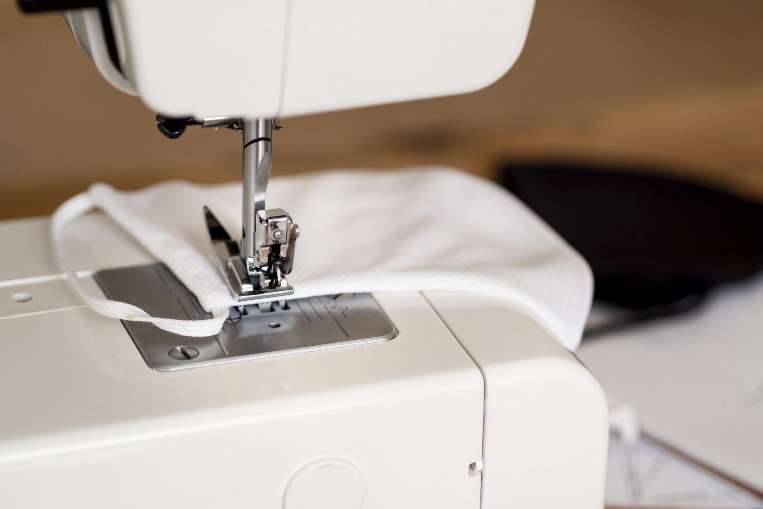 Close-up of a sewing machine stitching a white fabric face mask with elastic ear loops.