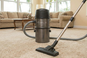 A person using a central vacuum powerhead attachment to deep clean a plush beige carpet in a sunlit living room.