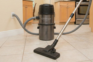 A powerful central vacuum system cleaning ceramic tile floors in a kitchen, showing the canister unit on the floor.